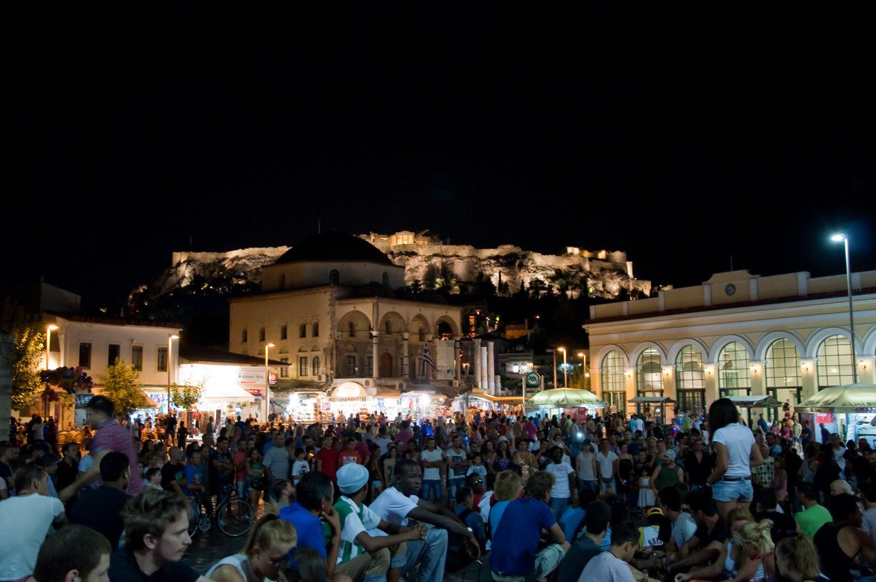 A vibrant night scene in Monastiraki, Athens, featuring a large crowd of people enjoying the lively atmosphere. The square is illuminated by streetlights and lights from surrounding buildings, including a mosque with a domed roof and a neoclassical building with arched windows. In the background, the Acropolis is dramatically lit against the night sky, creating a stunning backdrop. The image captures the energetic nightlife and cultural richness of the area.